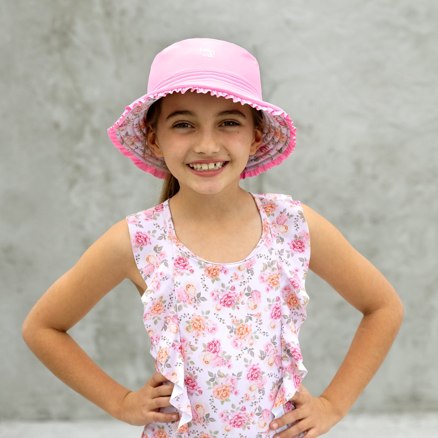 Young girl wearing a pink sun hat and floral sleeveless top against a gray background