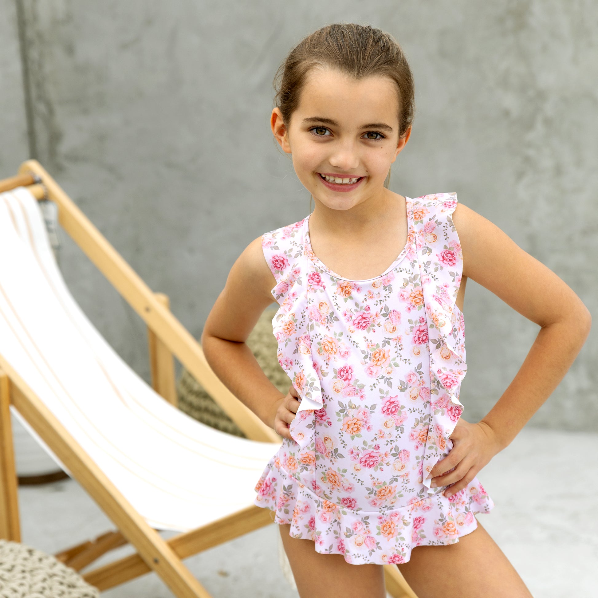 Young girl wearing a floral swimsuit standing in front of a wooden chair.