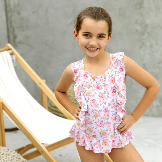 Young girl wearing a floral swimsuit standing in front of a wooden chair.