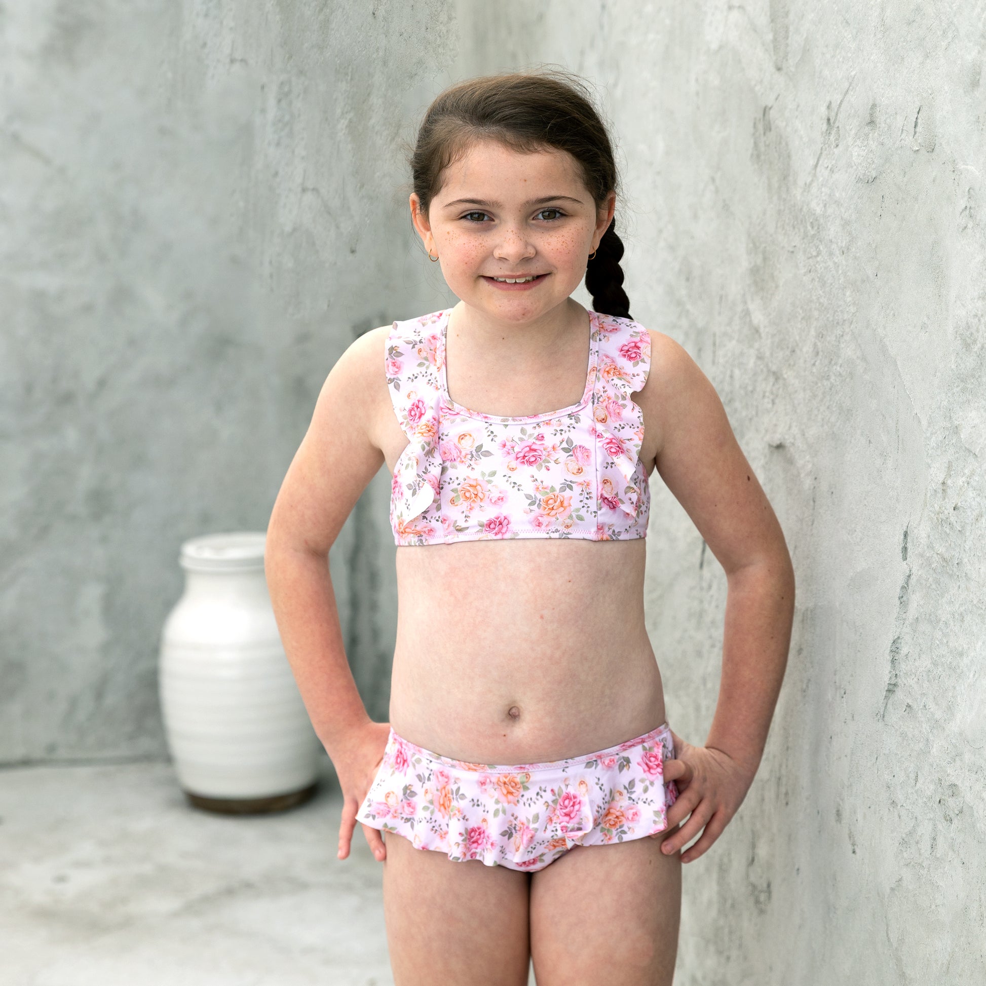 Young girl wearing a floral two-piece swimsuit against a gray concrete wall.