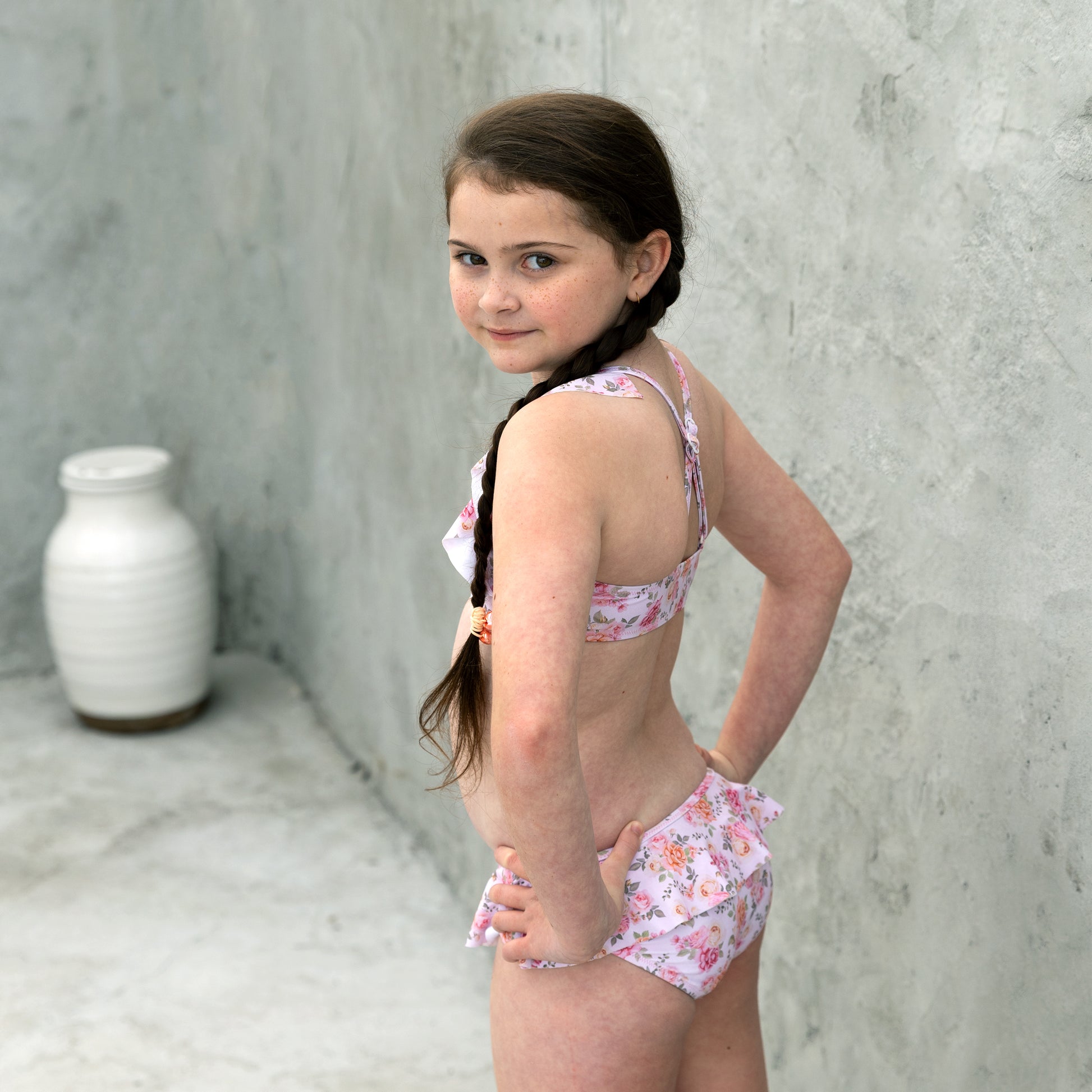 Young girl wearing a floral swimsuit against a concrete wall.