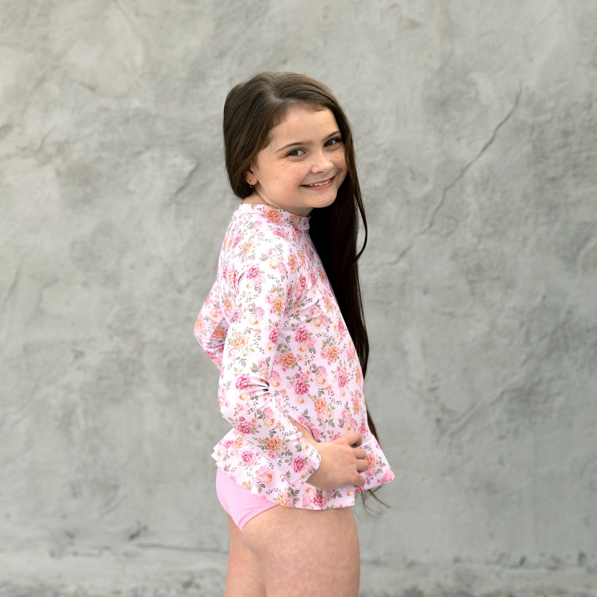 Young girl wearing a floral rash guard and pink shorts against a gray background