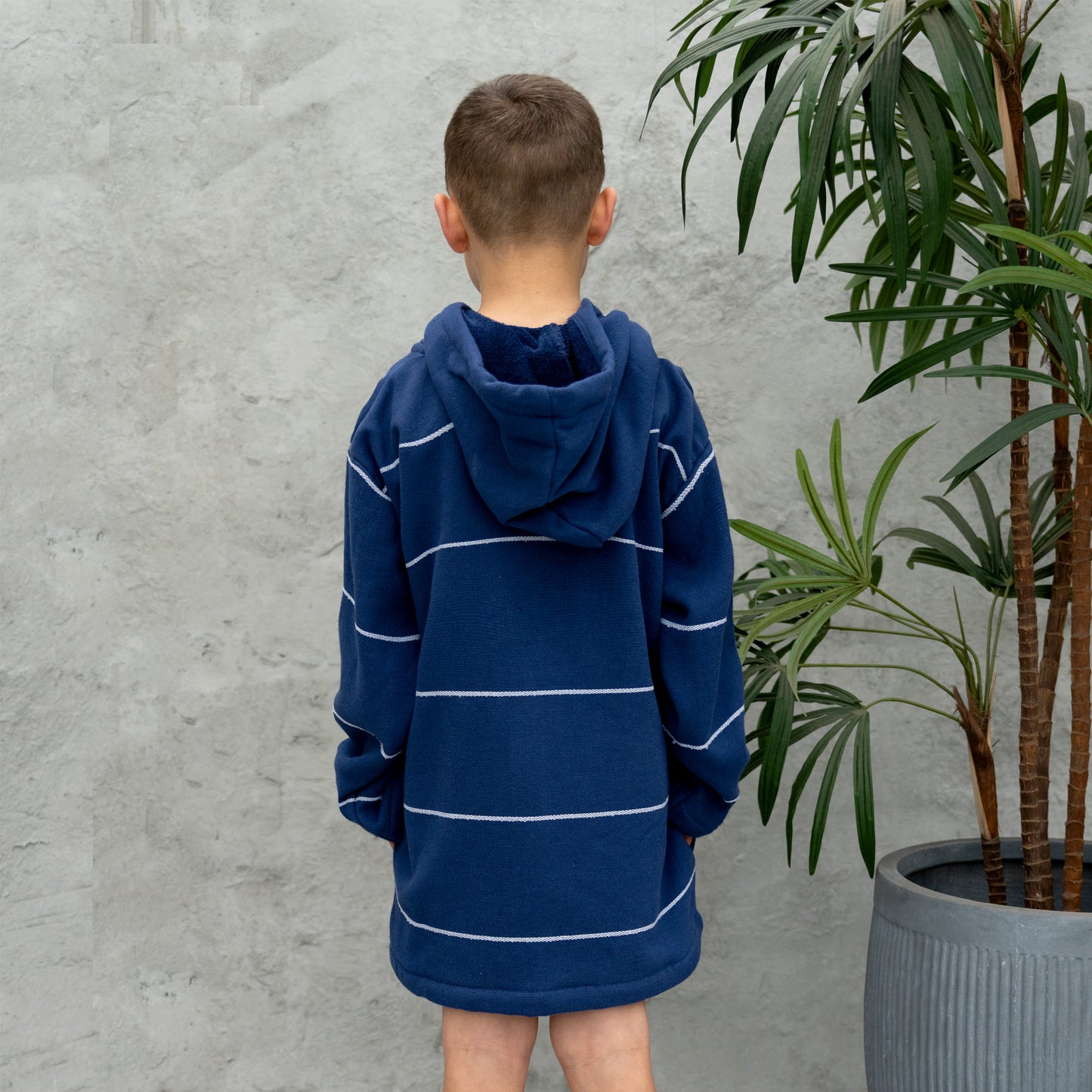 Child wearing a blue striped hoodie standing against a gray wall with a plant in the background