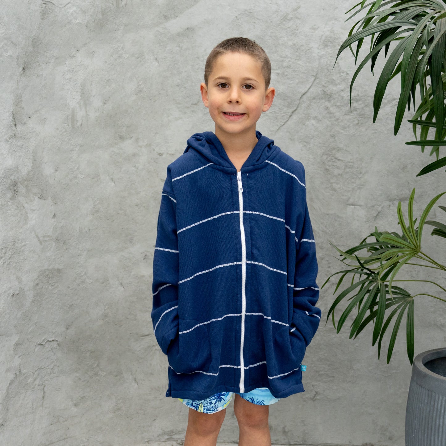 Child wearing a blue striped hoodie standing against a gray wall with a plant.
