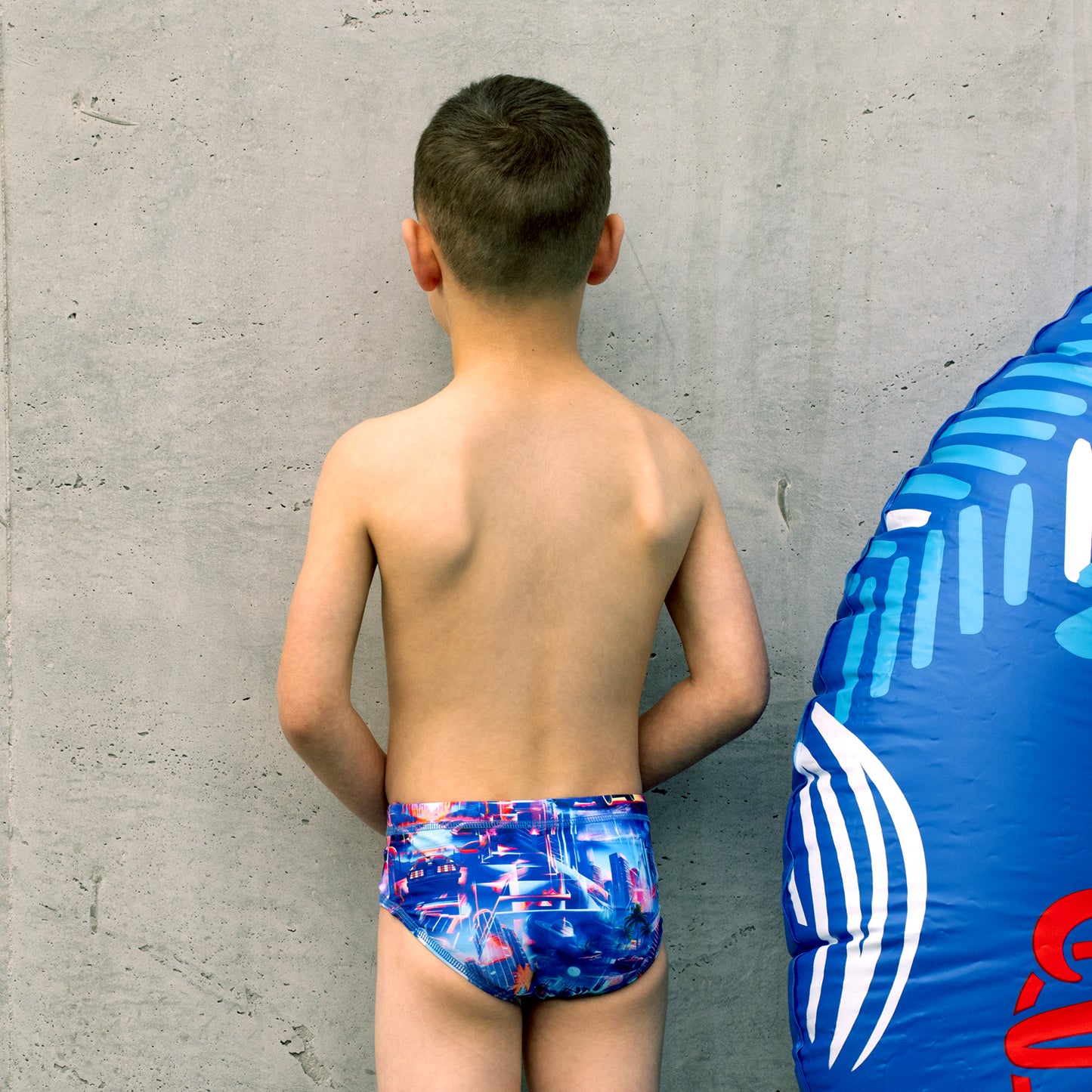 Young boy wearing colorful boys swim briefs standing against a concrete wall with a blue inflatable ring on the ground.