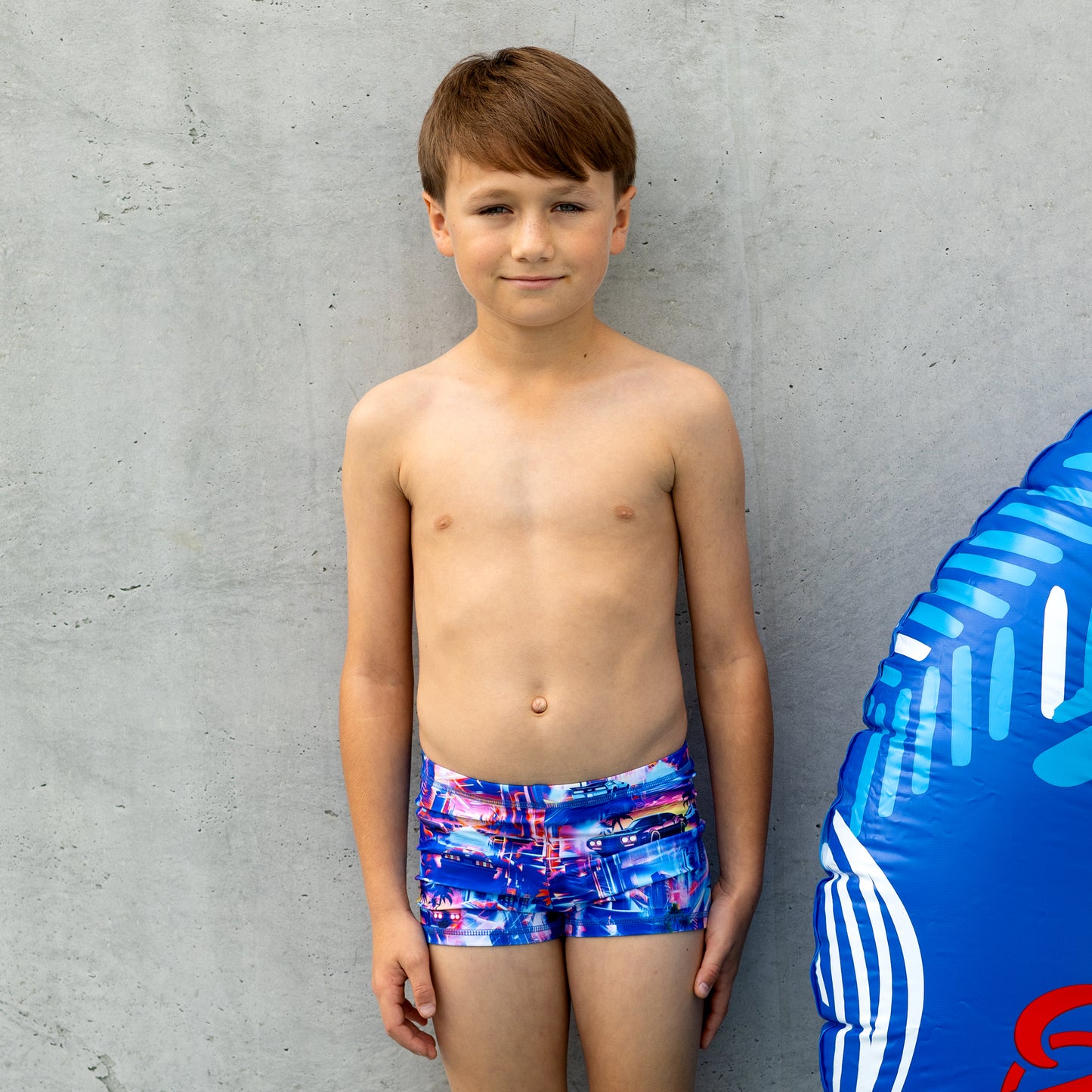 Young boy wearing colorful boy swim trunks standing next to a blue inflatable ring against a gray wall.