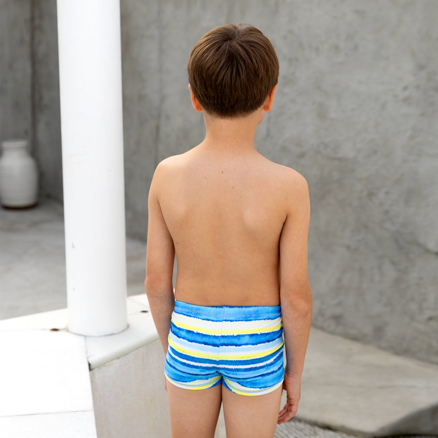 Young boy wearing blue boys swim trunks with yellow stripes standing outdoors.