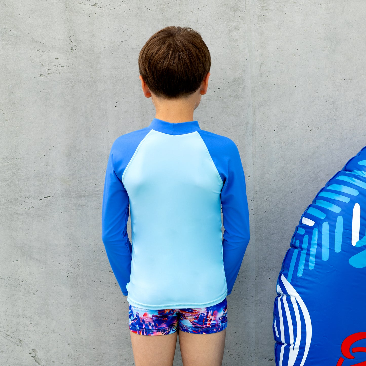 Child wearing a blue and light blue long-sleeve salty ink boy rashie with colorful graphics and shorts, standing next to an inflatable toy against a gray wall.
