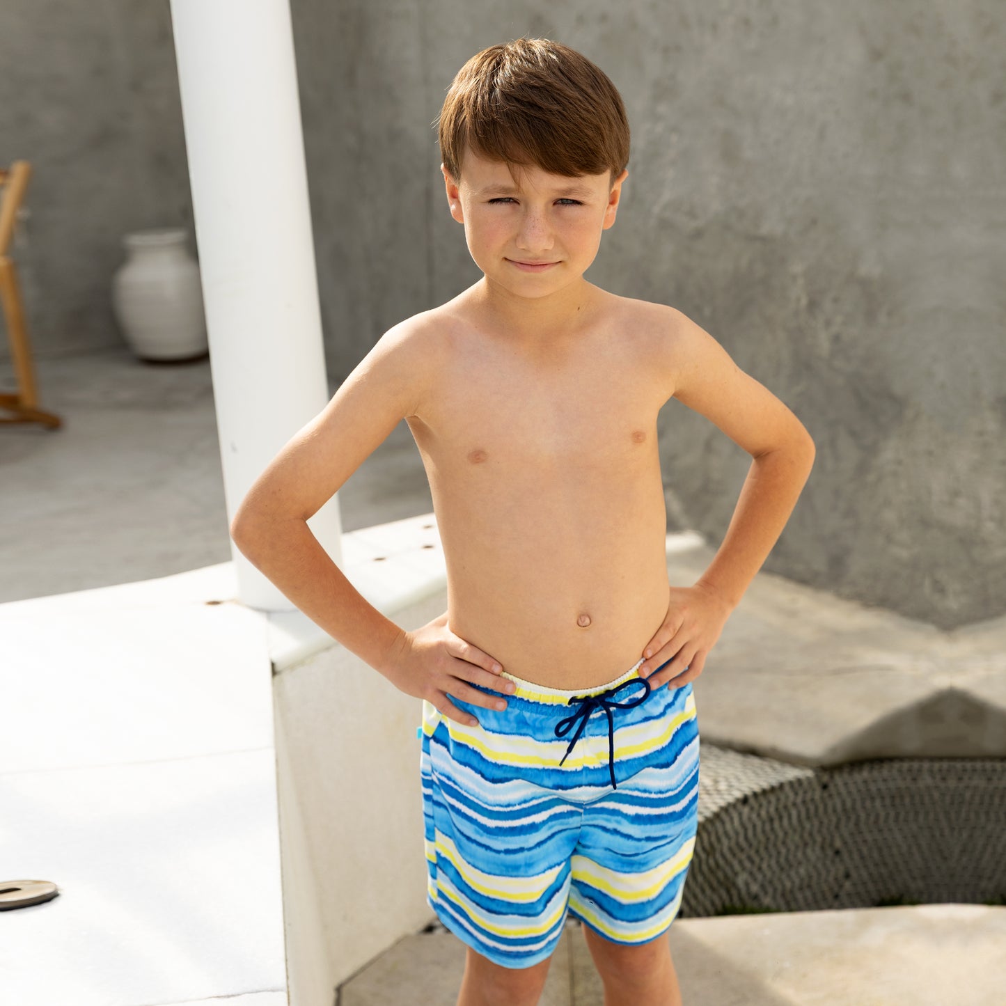Young boy wearing blue and yellow striped boys swim board shorts standing on a white surface.