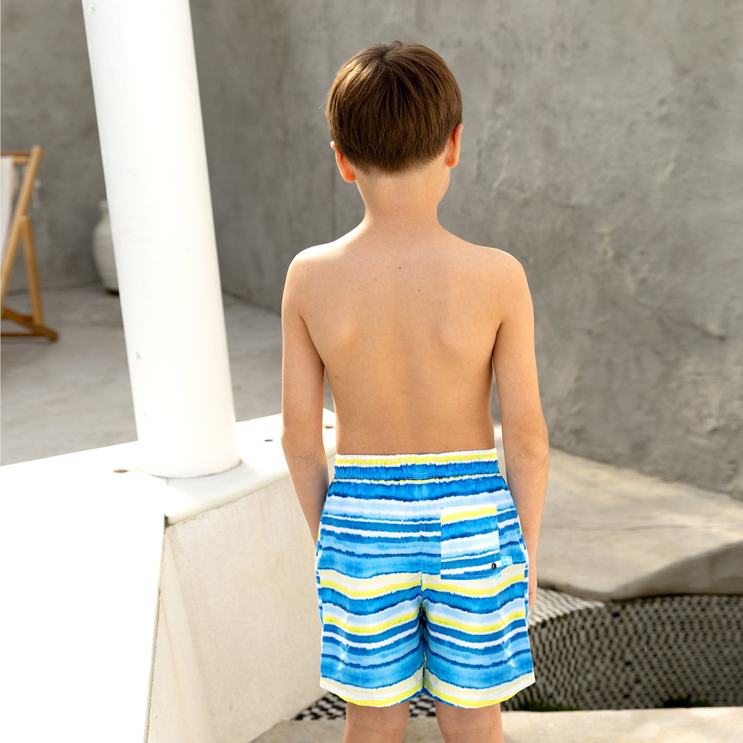 Young boy wearing blue and yellow striped boys swim board shorts standing on a white surface.