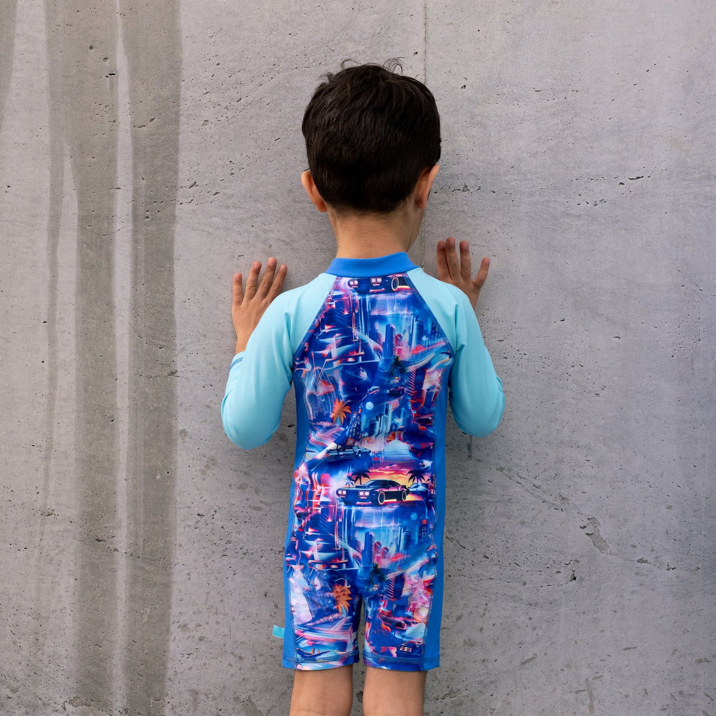 Child wearing a colorful boys all in one swimsuit with blue sleeves against a gray concrete wall