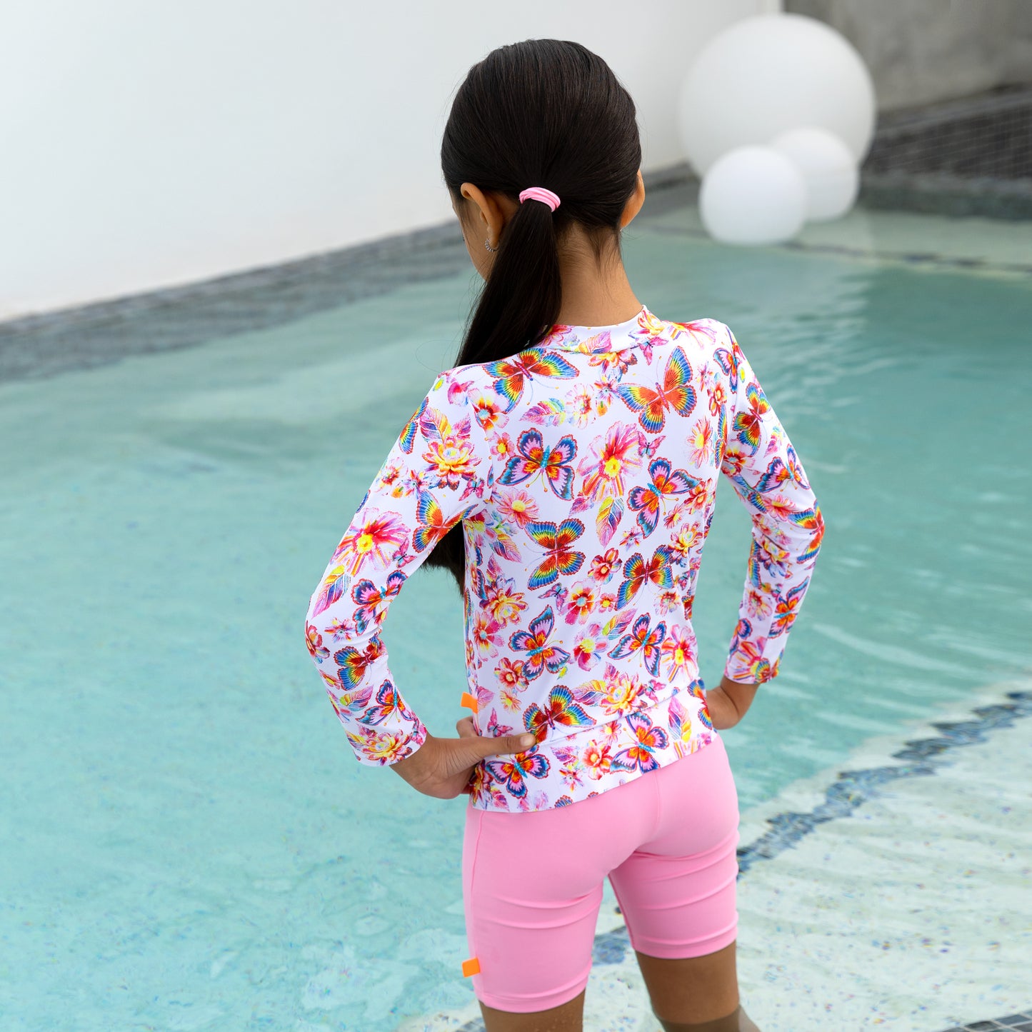 Child wearing a colorful butterfly-patterned long-sleeve shirt and pink shorts by a pool.