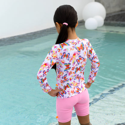 Child wearing a colorful butterfly-patterned long-sleeve shirt and pink shorts by a pool.