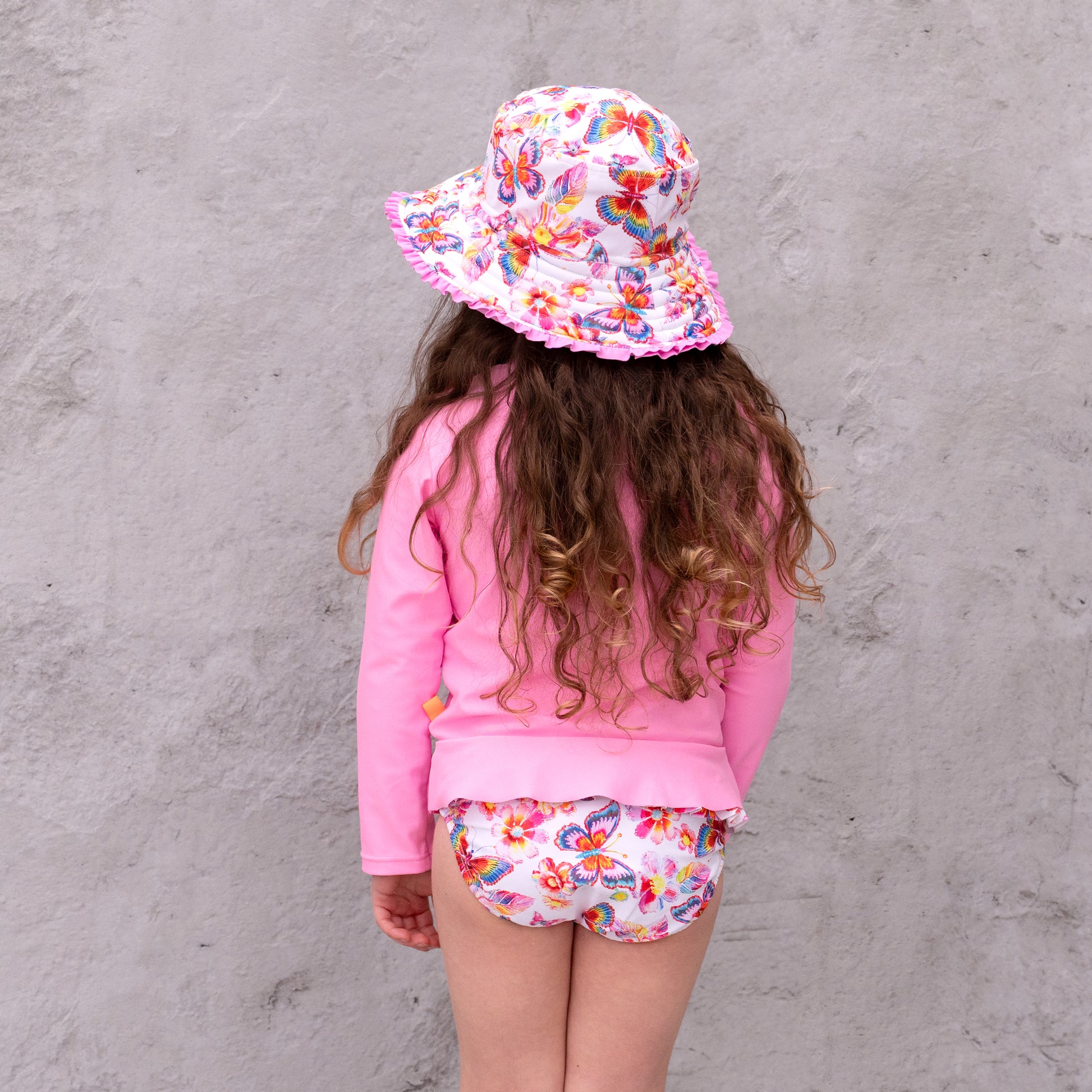 Child wearing a pink outfit with floral hat and shorts against a gray wall