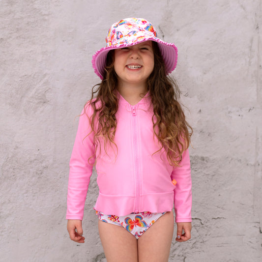Young girl wearing a pink UV protective outfit with a colorful hat against a gray wall.