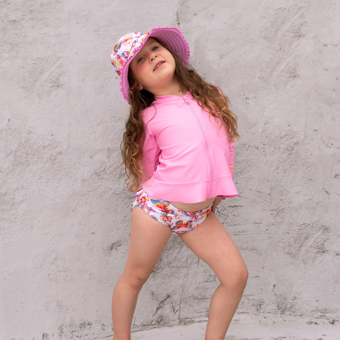 Young girl in pink UV protective outfit with floral hat and swim diapers against a gray concrete wall.