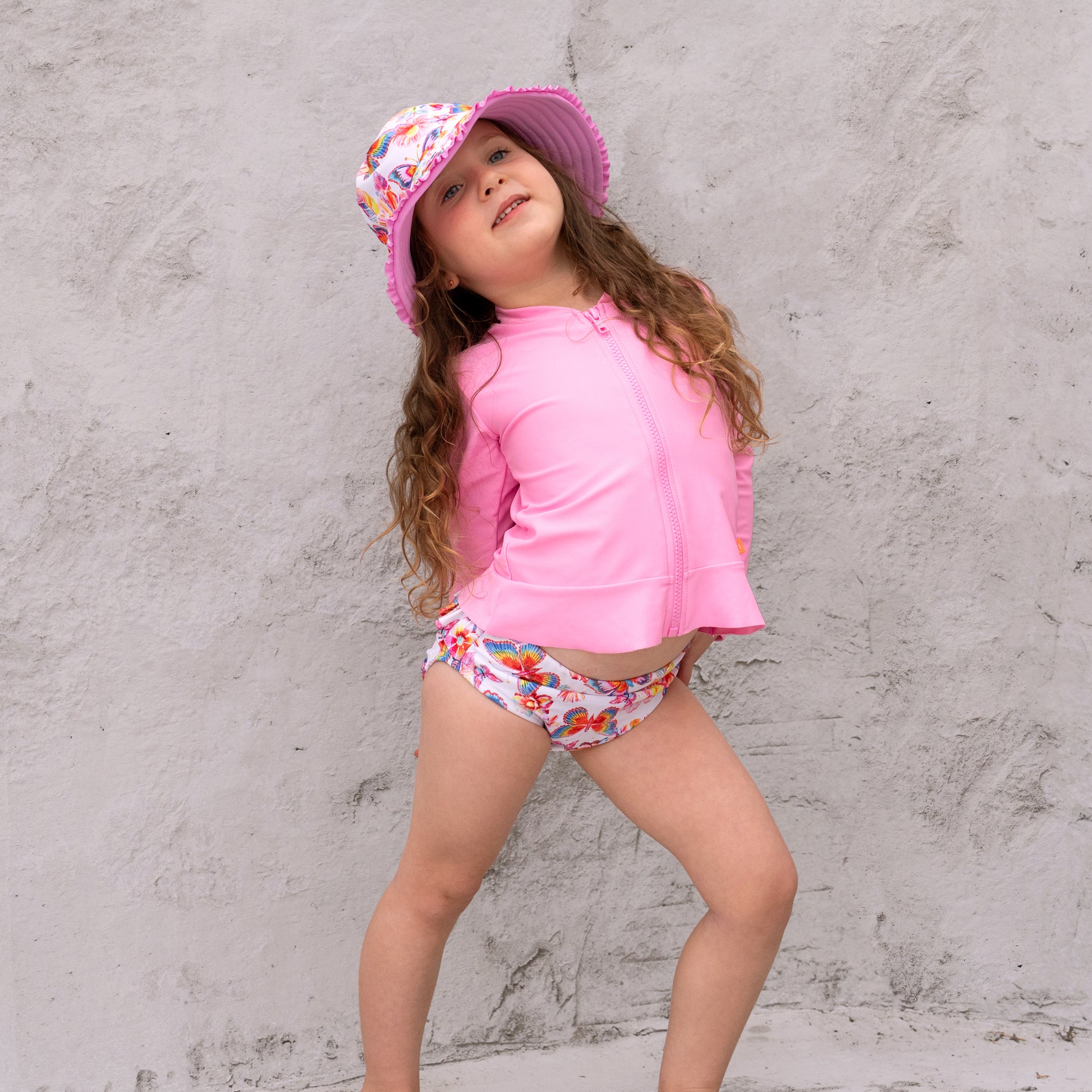 Young girl in pink UV protective outfit with floral hat and swim diapers against a gray concrete wall.