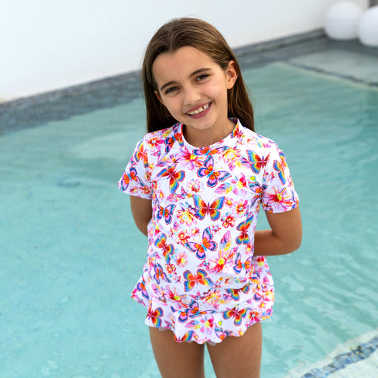 Young girl in a colorful butterfly swimsuit standing by a pool.