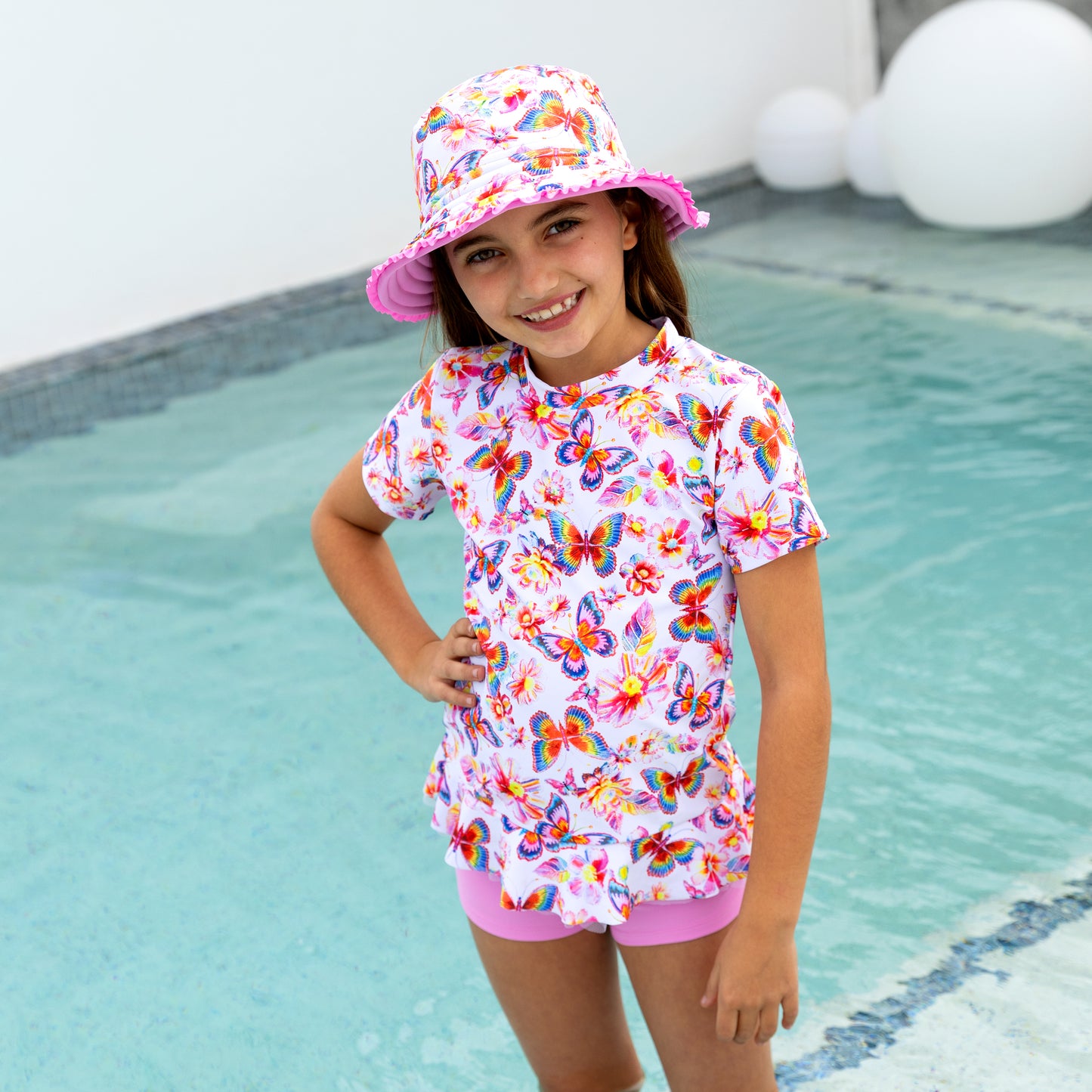 Child wearing a colorful butterfly-patterned swimsuit and hat by a pool.