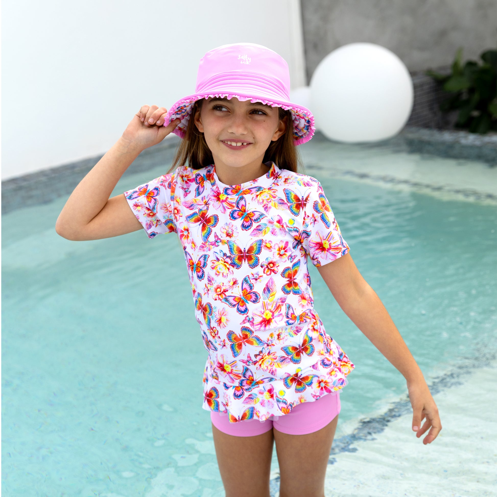 Young girl in a butterfly-patterned swimsuit and pink hat by a pool.