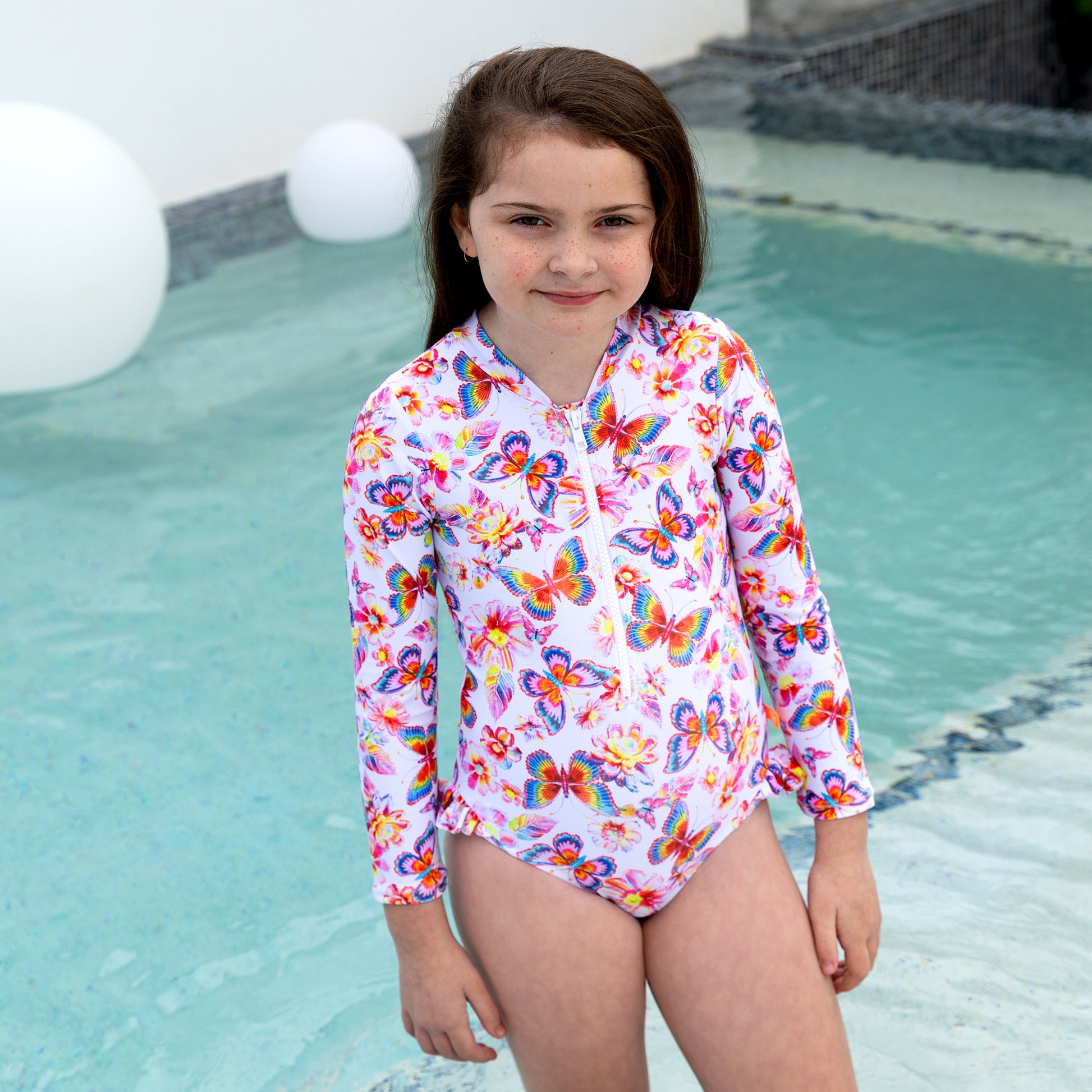 Young girl in a colorful butterfly-patterned swimsuit standing by a pool.