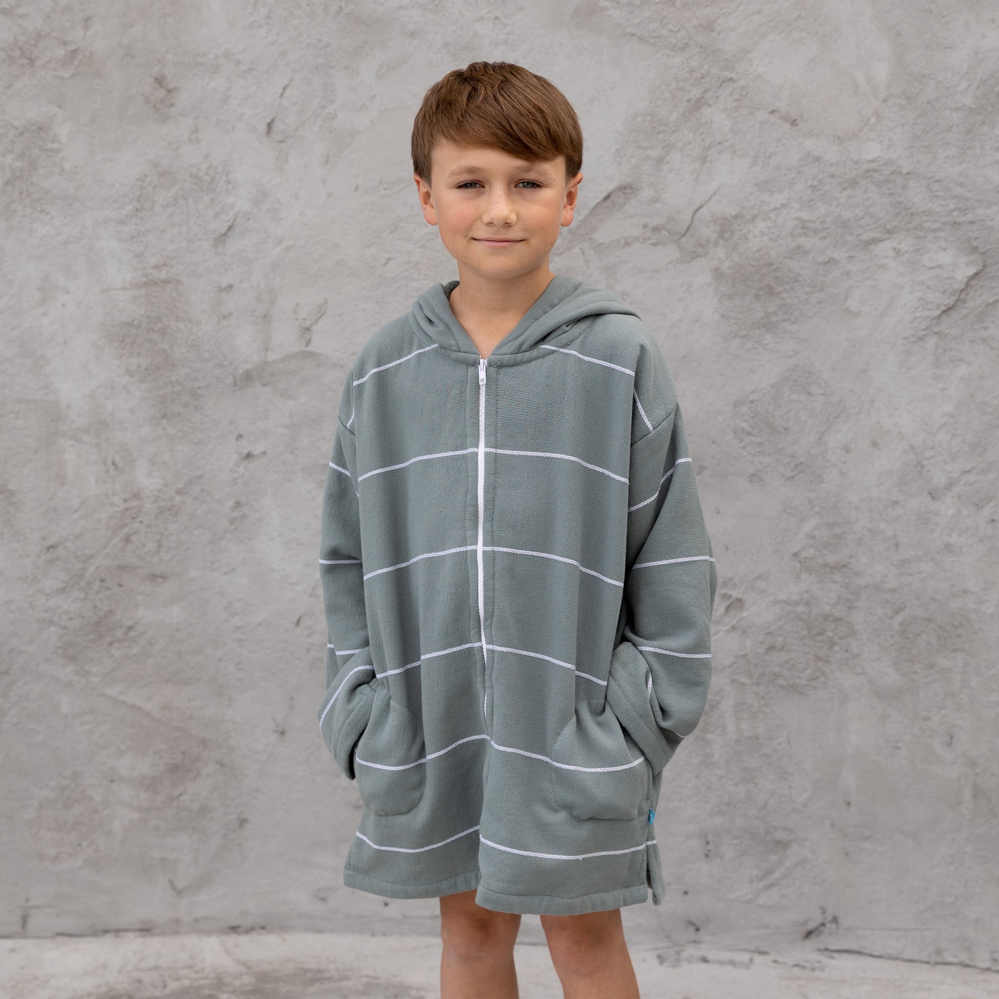 Child wearing a gray hooded towel with white stripes against a gray concrete wall.