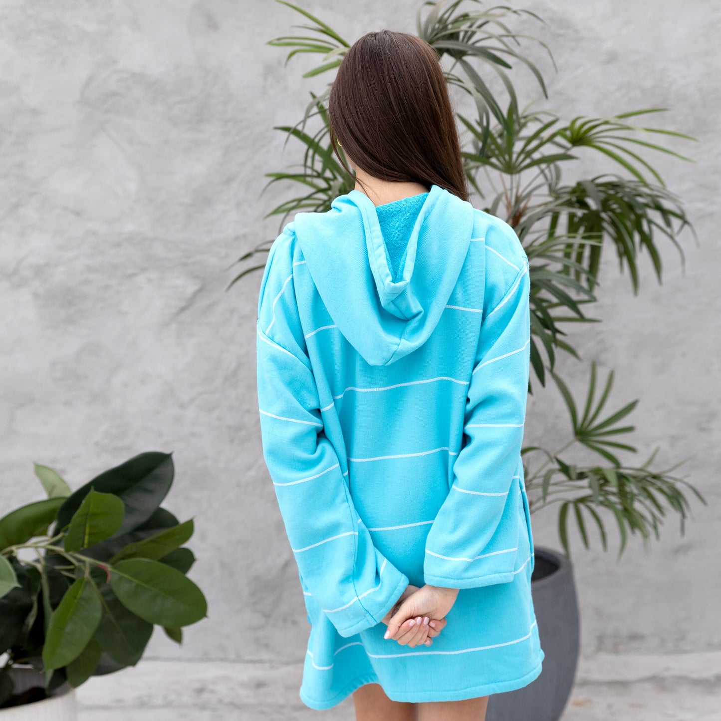 Person wearing a blue hooded towel against a gray wall with plants.