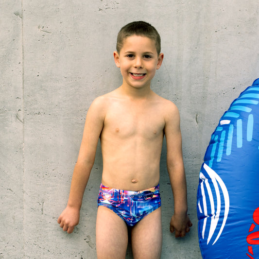 Young boy wearing colorful boys swim briefs standing against a concrete wall with a blue inflatable ring on the ground.