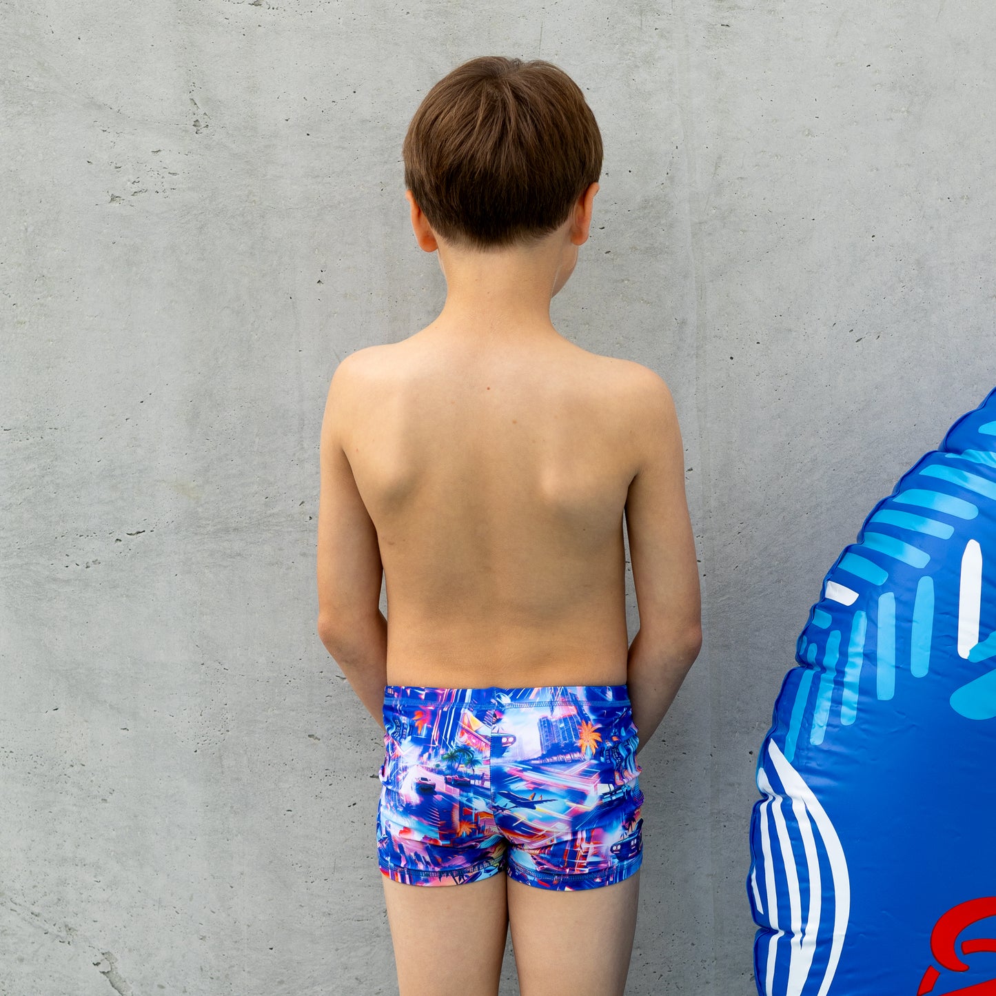Young boy wearing colorful boy swim trunks standing next to a blue inflatable ring against a gray wall.