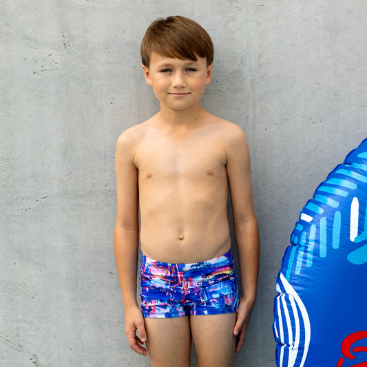 Young boy wearing colorful boy swim trunks standing next to a blue inflatable ring against a gray wall.