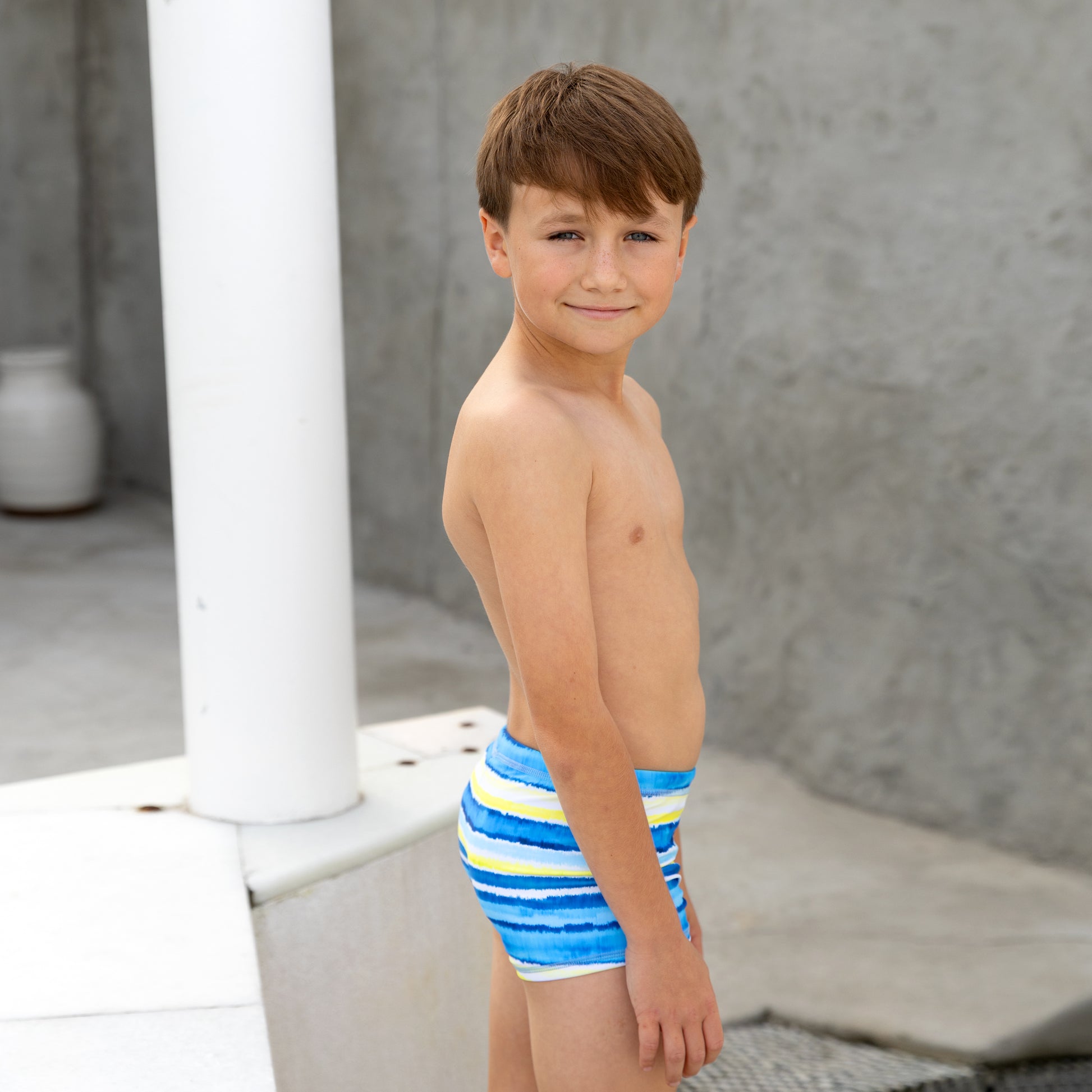 Young boy wearing blue boys swim trunks with yellow stripes standing outdoors.