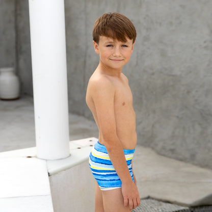 Young boy wearing blue boys swim trunks with yellow stripes standing outdoors.
