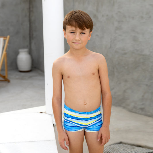 Young boy wearing blue boys swim trunks with yellow stripes standing outdoors.