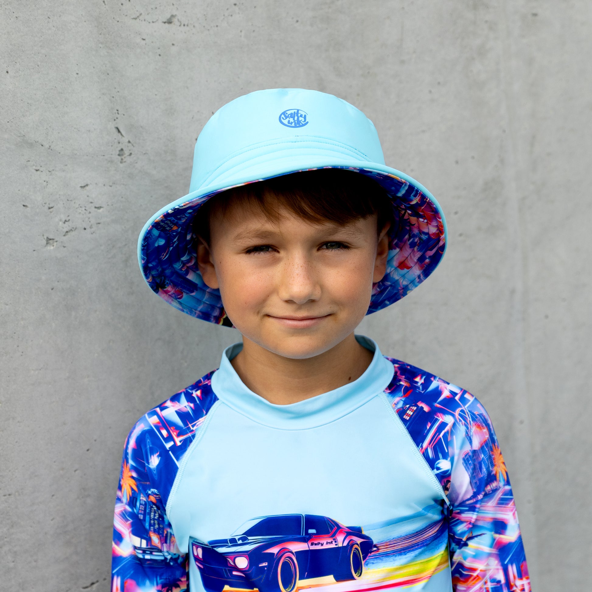 Child wearing a colorful salty ink bucket swim hat and rash guard against a gray wall