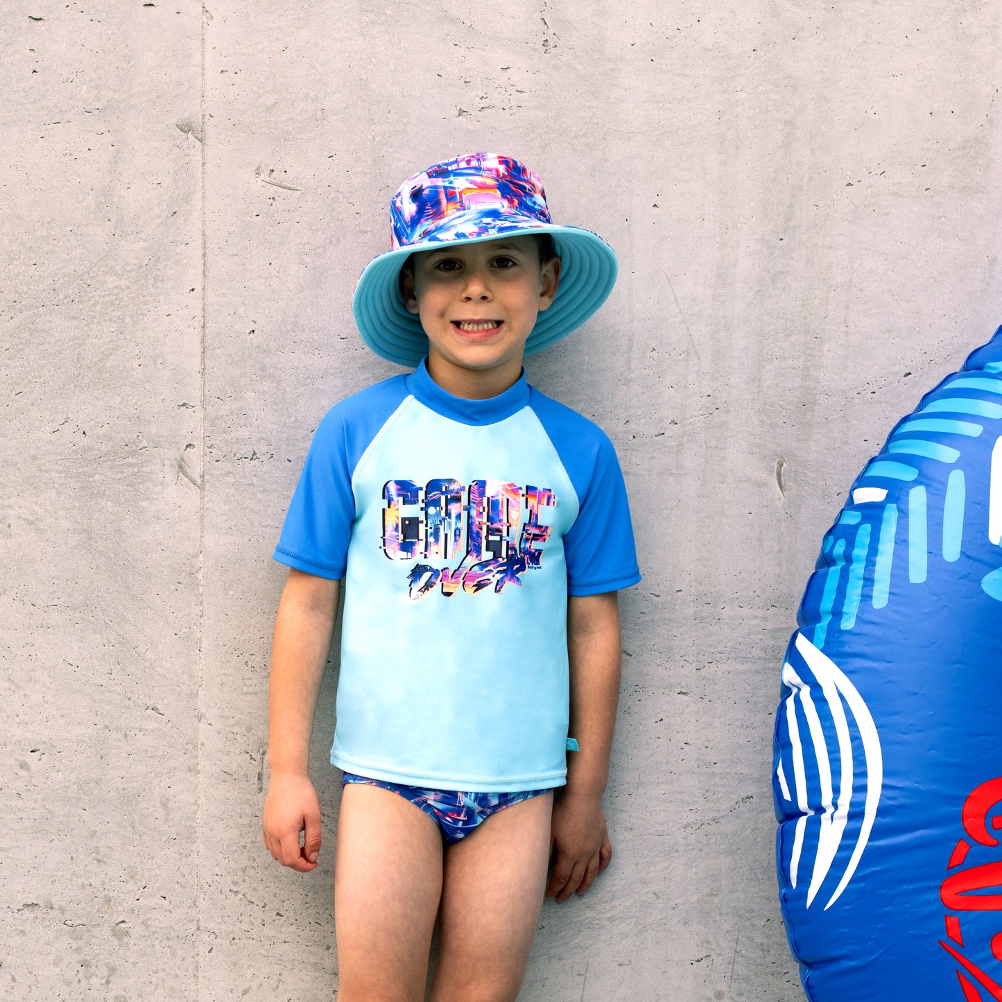 Child wearing a colorful boys salty ink bucket swim hat and rash guard against a gray wall