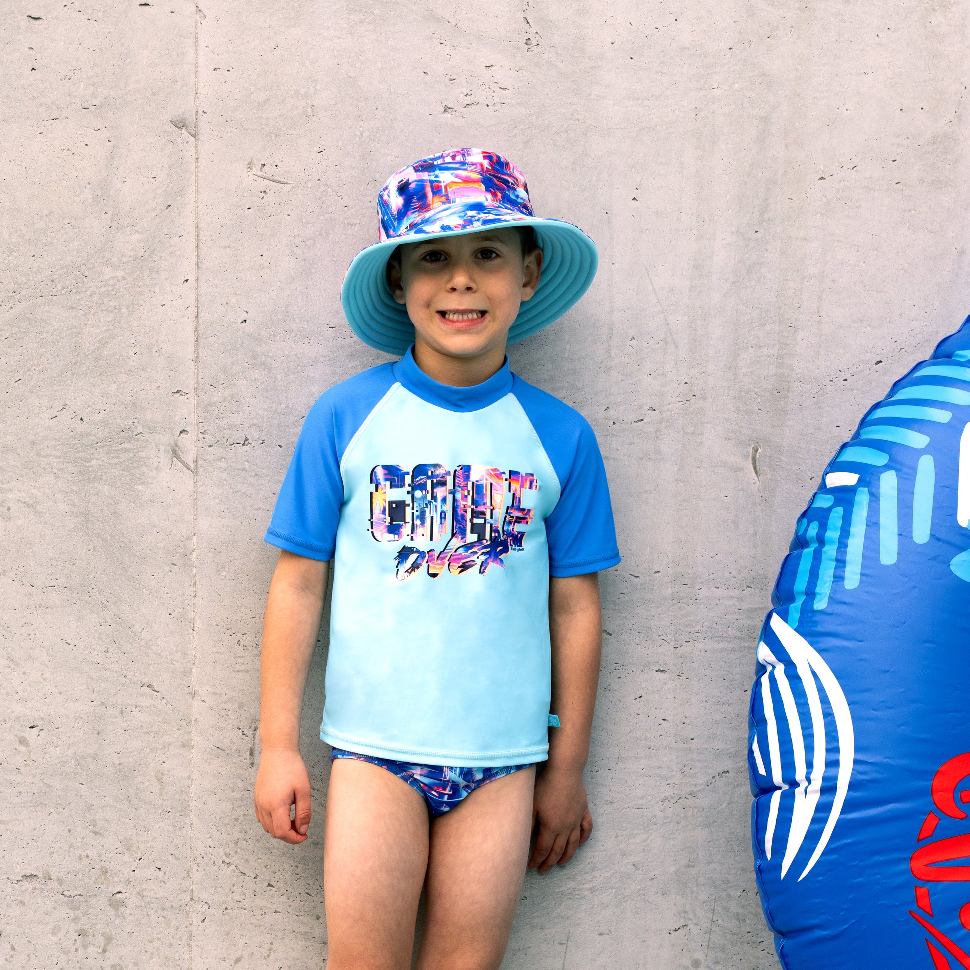 Child wearing a colorful boys salty ink bucket swim hat and rash guard against a gray wall