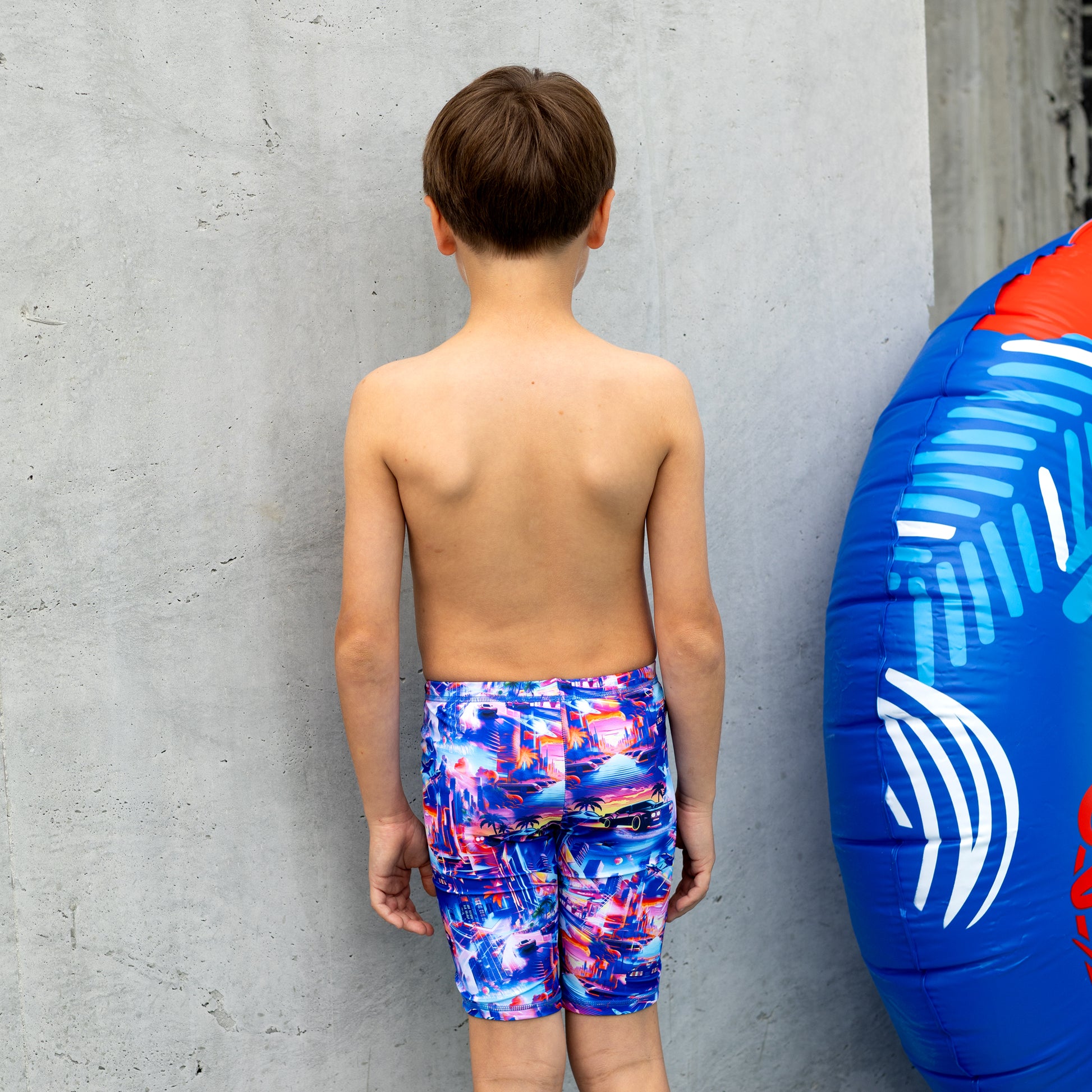 Young boy in colorful boy swim trunks standing next to a blue and red inflatable tube against a gray concrete wall.