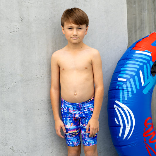Young boy in colorful boy swim trunks standing next to a blue and red inflatable tube against a gray concrete wall.