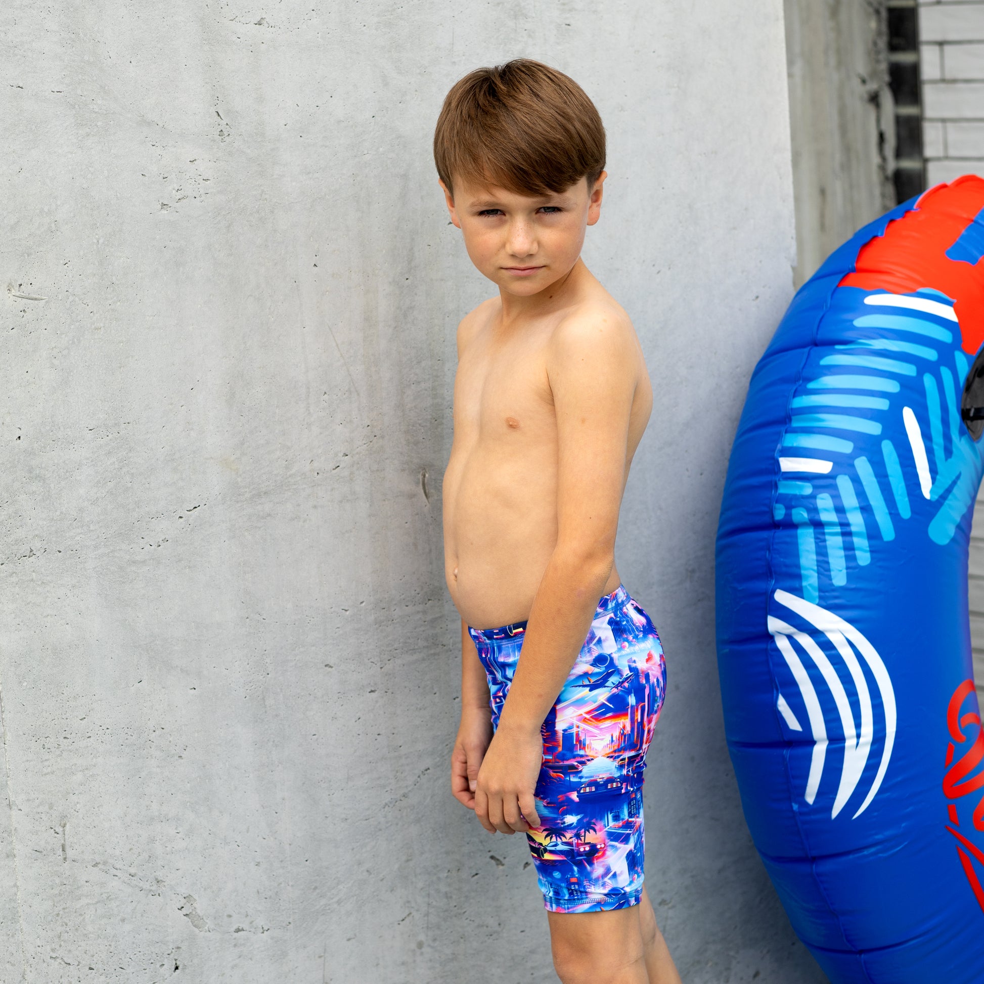 Young boy in colorful boy swim trunks standing next to a blue and red inflatable tube against a gray concrete wall.