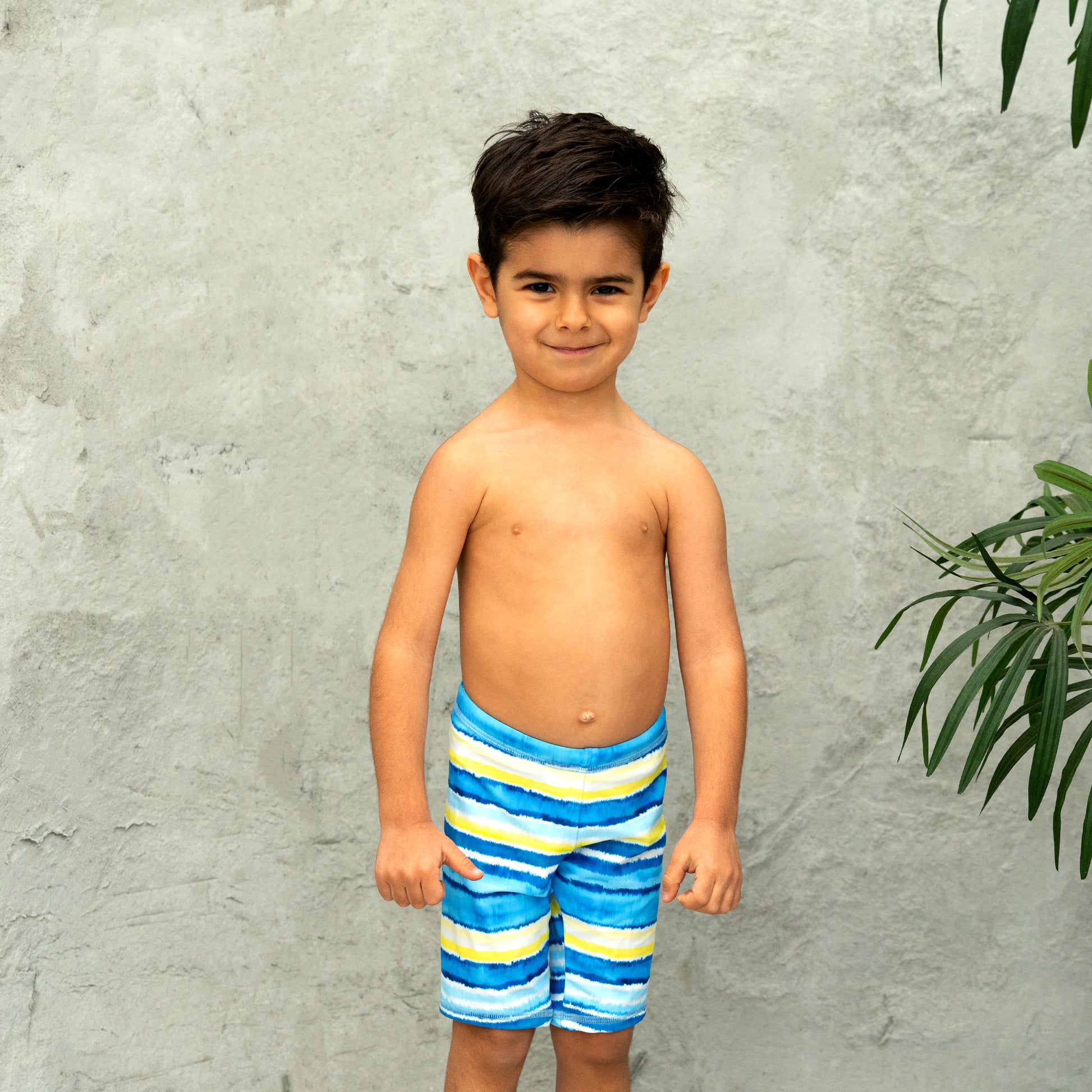 Young boy wearing blue and yellow striped swim shorts standing against a gray wall.