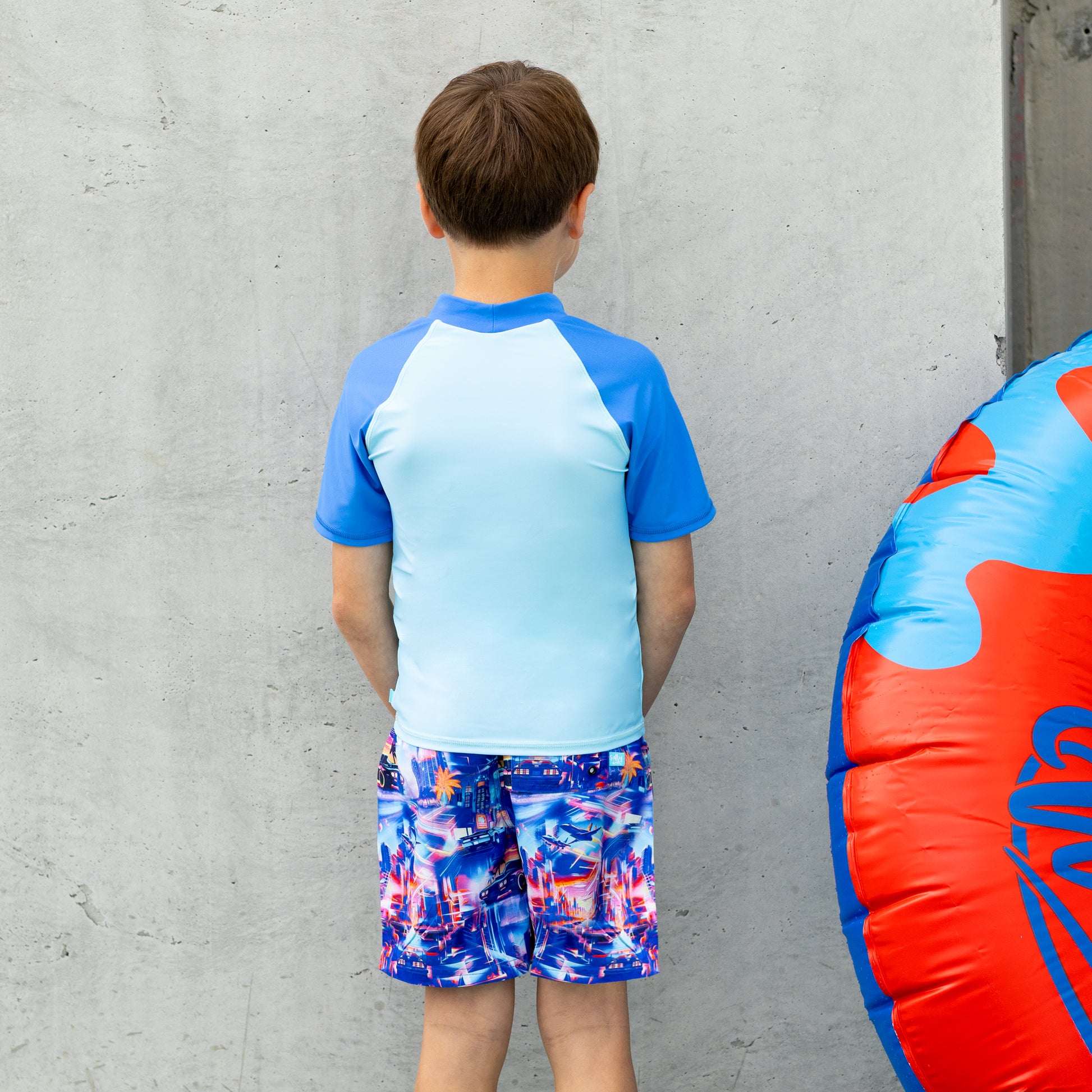 Child wearing a salty ink blue and multicolor boys rashie with matching shorts, standing against a gray wall.