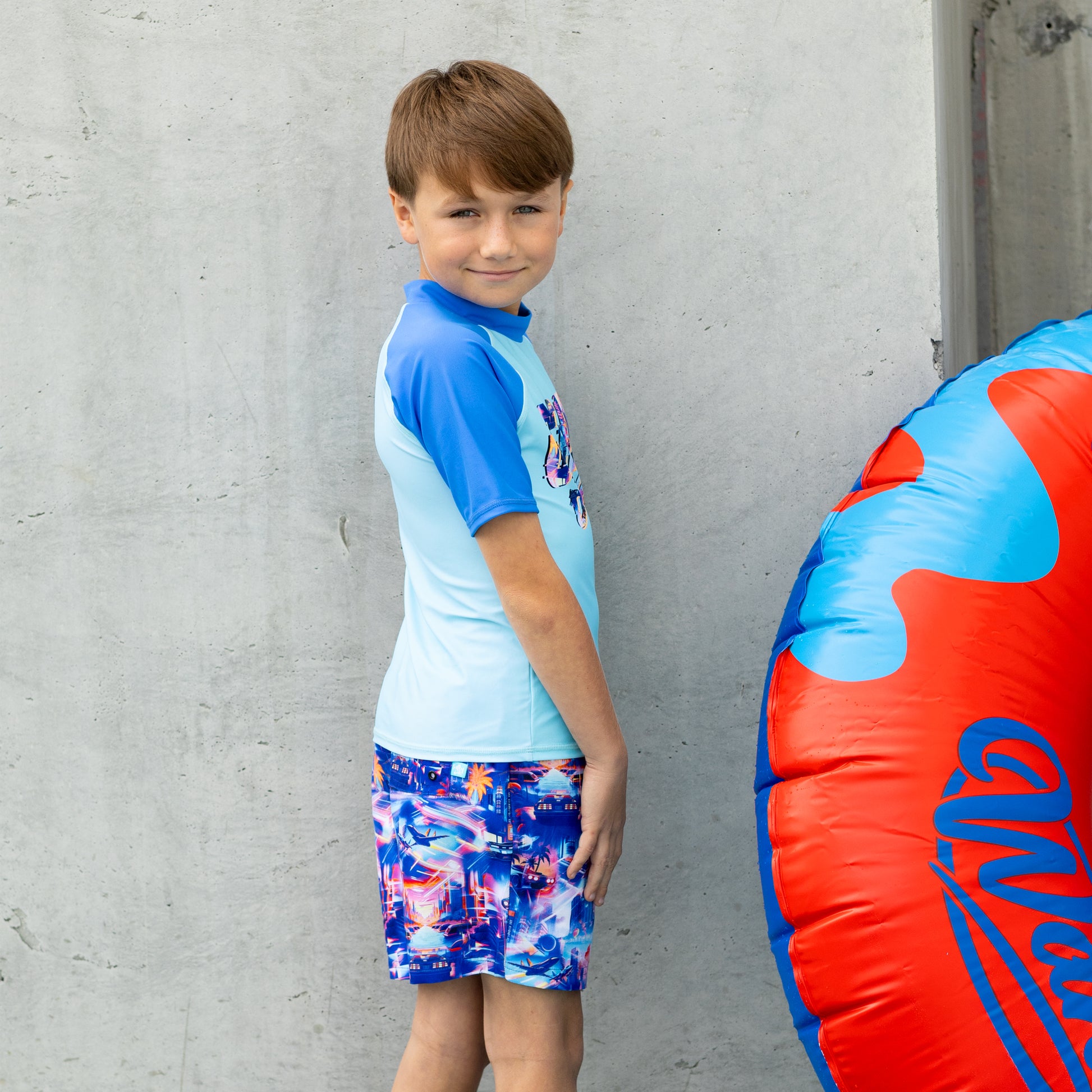 Child wearing a salty ink blue and multicolor boys rashie with matching shorts, standing against a gray wall.