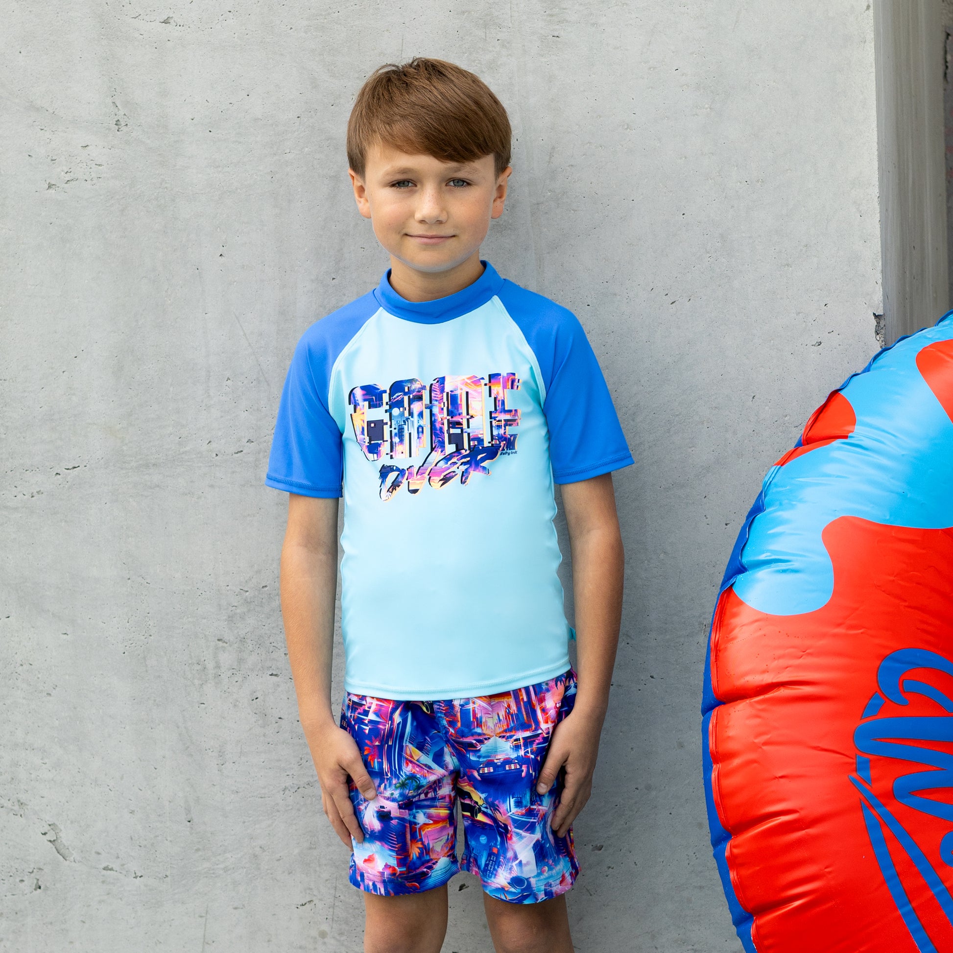Child wearing a salty ink blue and multicolor boys rashie with matching shorts, standing against a gray wall.