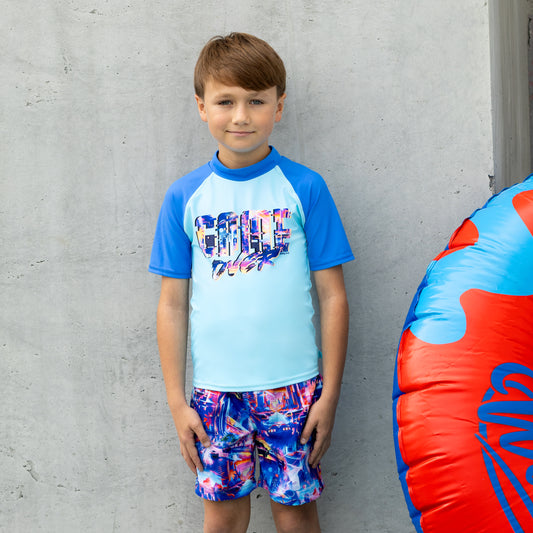 Child wearing a salty ink blue and multicolor boys rashie with matching shorts, standing against a gray wall.