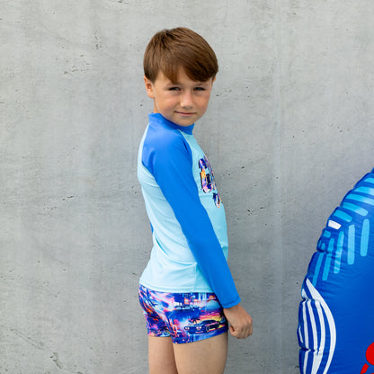 Child wearing a blue and light blue long-sleeve salty ink boy rashie with colorful graphics and shorts, standing next to an inflatable toy against a gray wall.