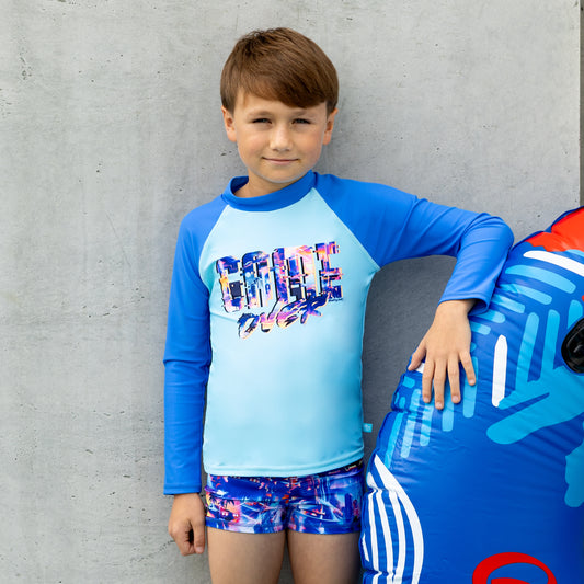 Child wearing a blue and light blue long-sleeve salty ink boy rashie with colorful graphics and shorts, standing next to an inflatable toy against a gray wall.