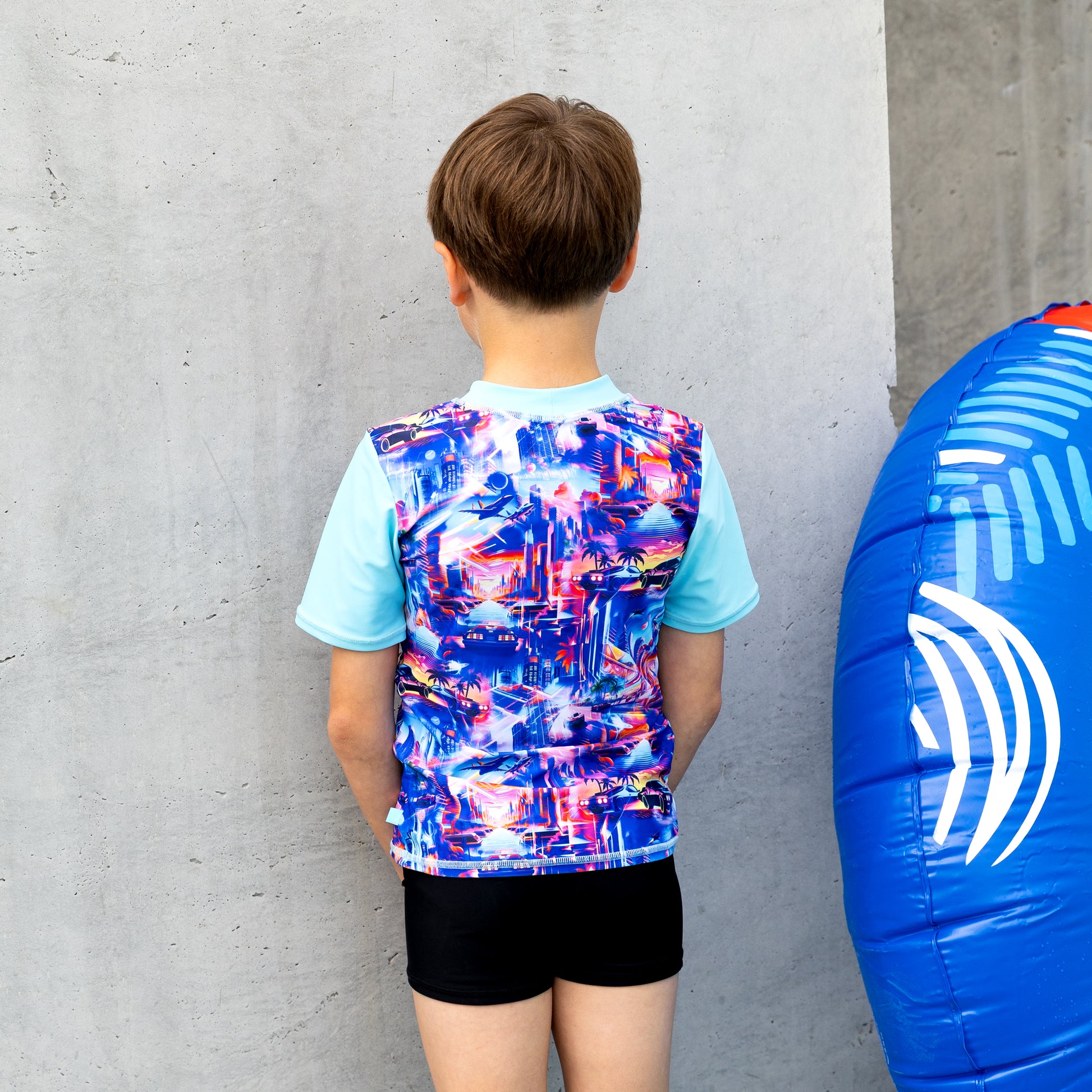Child wearing a boy rashie with a blue and white inflatable ring next to them against a concrete wall.