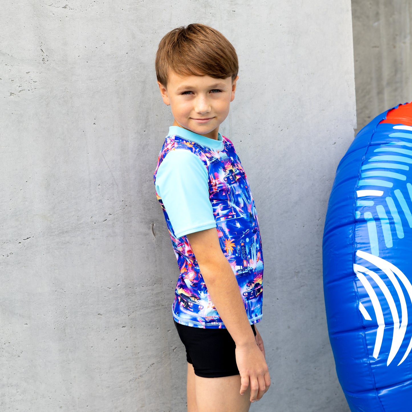 Child wearing a boy rashie with a blue and white inflatable ring next to them against a concrete wall.