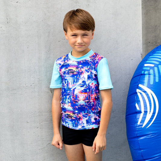 Child wearing a boy rashie with a blue and white inflatable ring next to them against a concrete wall.