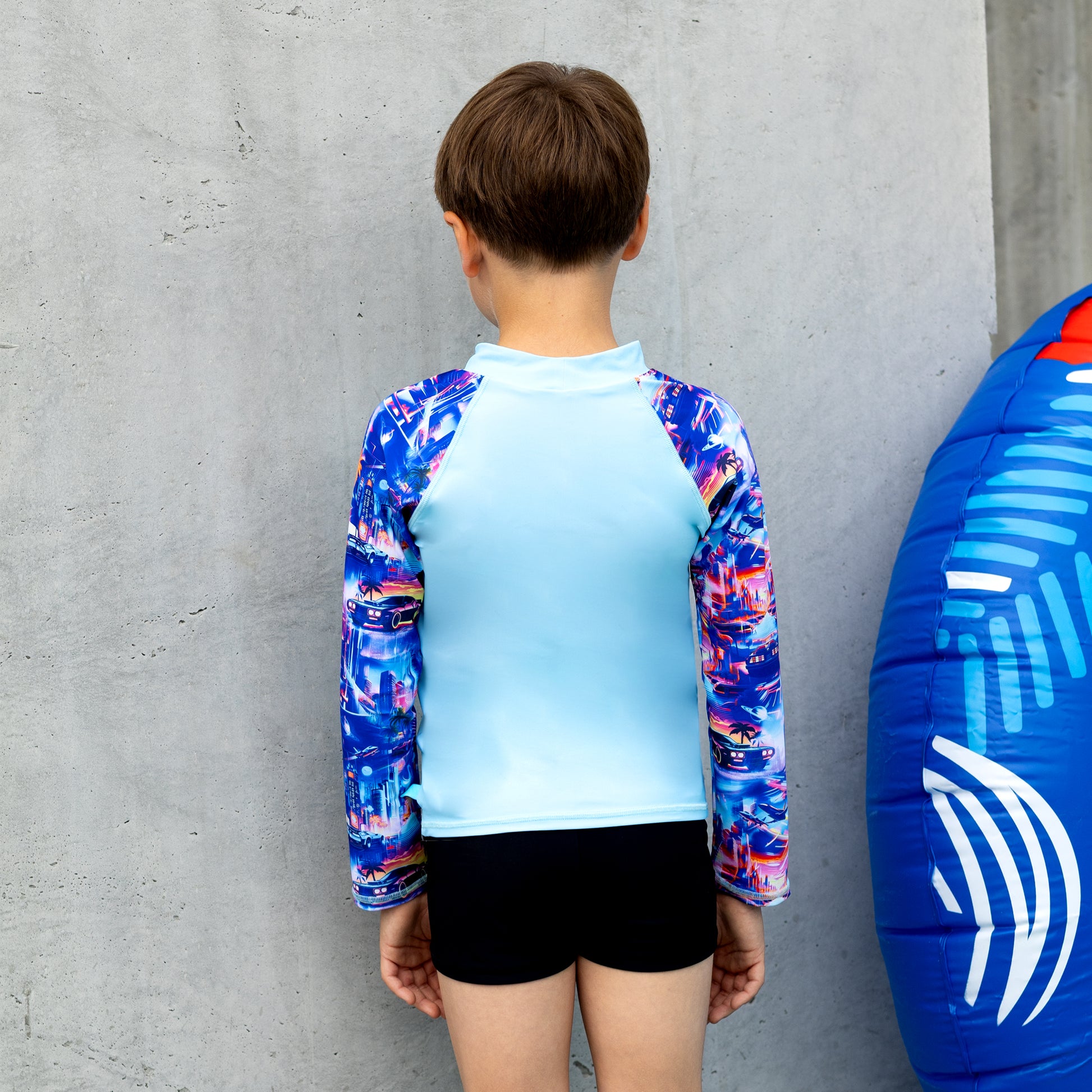 Child wearing a boy long sleeve  rashie with a blue and white inflatable ring next to them against a concrete wall.
